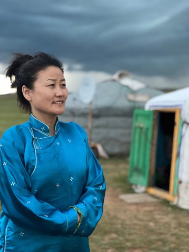 Wohin schaut Sie? Frau in blauer traditioneller Kleidung vor einer Jurte unter bewölkten Himmel.